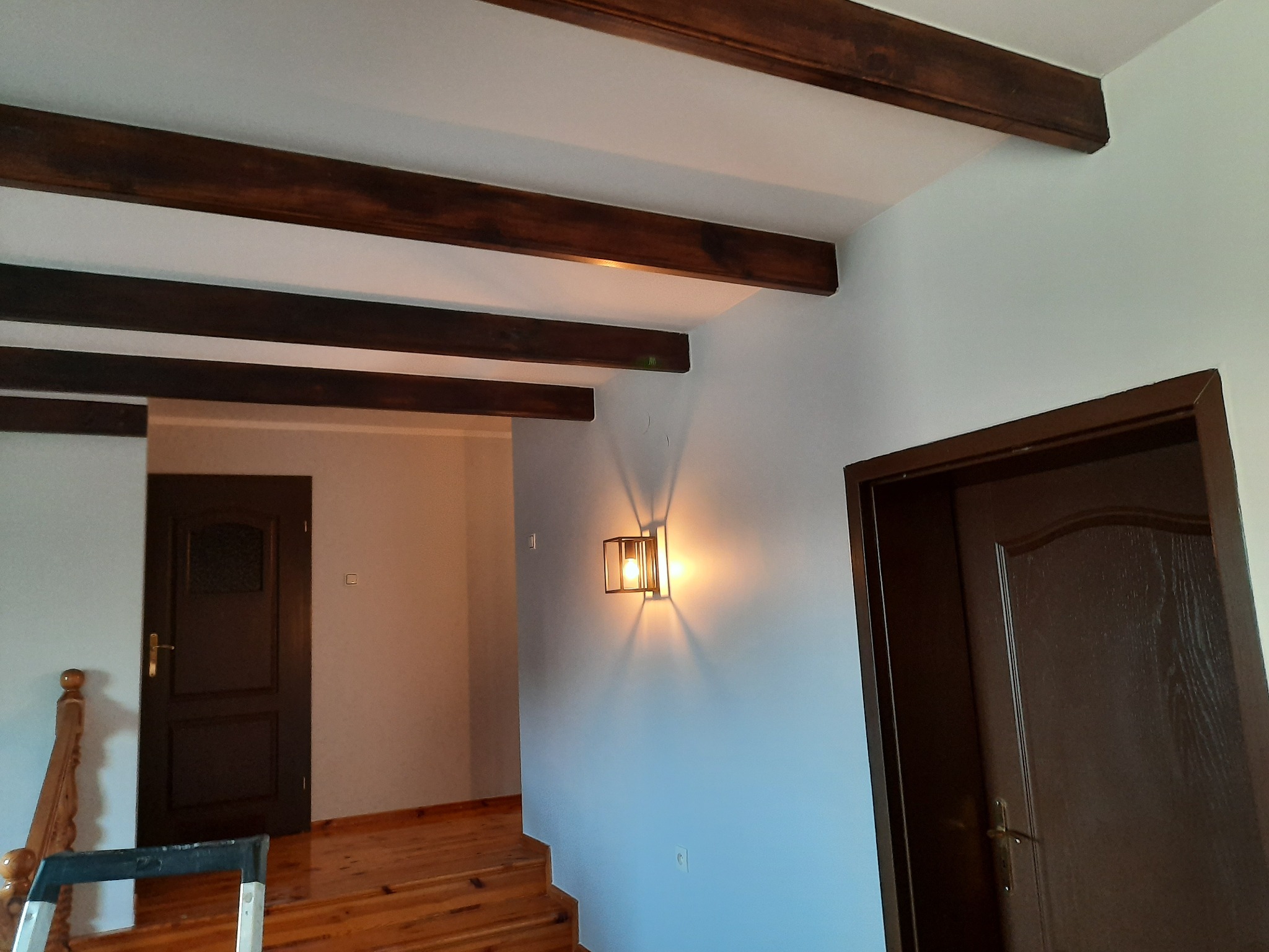 Wnętrze domu z drewnianymi belkami stropowymi, oświetleniem ściennym w stylu industrialnym i drewnianymi schodami oraz drzwiami.