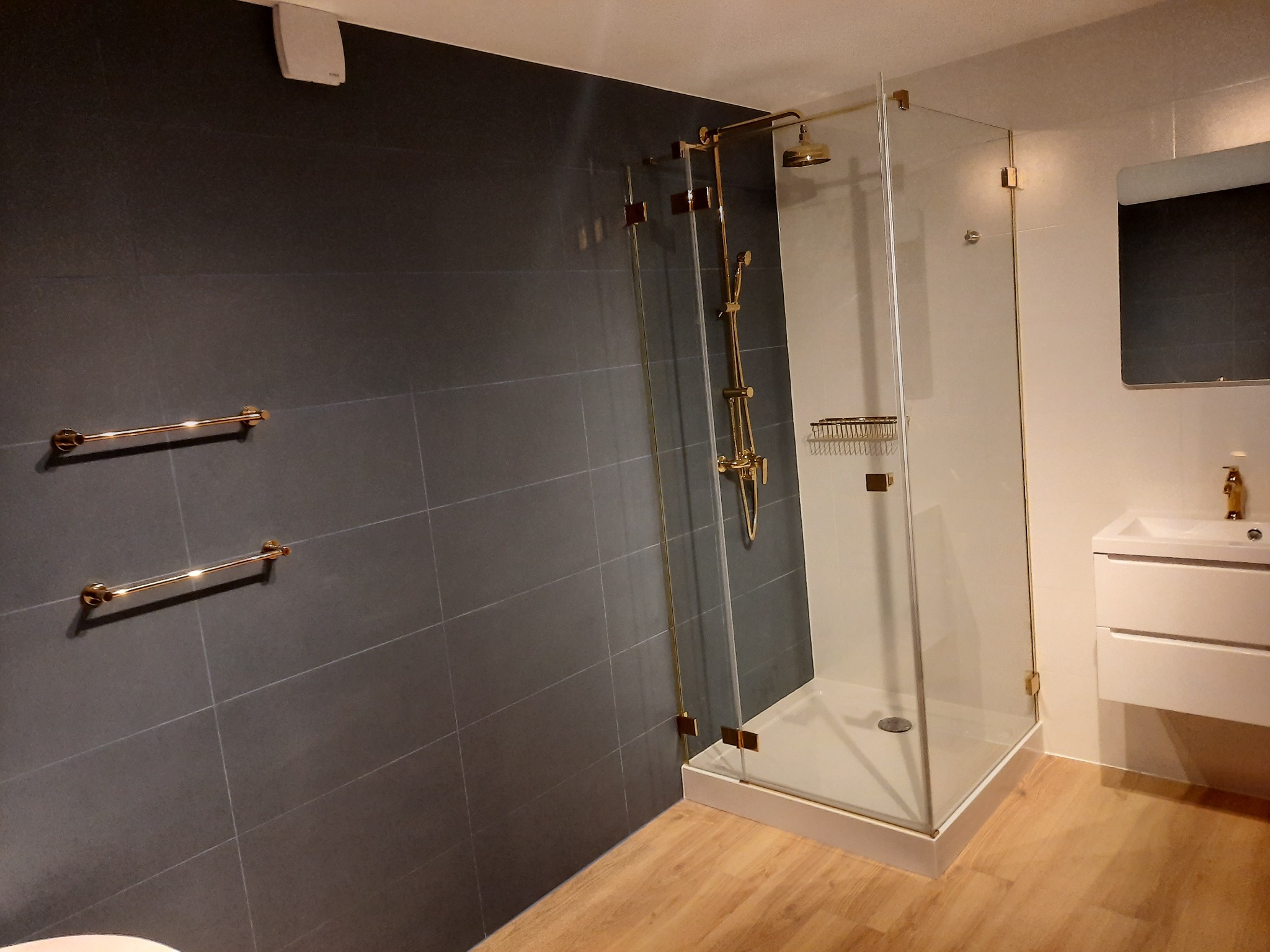 Nowoczesna łazienka z kabiną prysznicową ze złotymi elementami, ciemnoszare płytki na ścianie, drewnopodobna podłoga i biała umywalka z szafką.