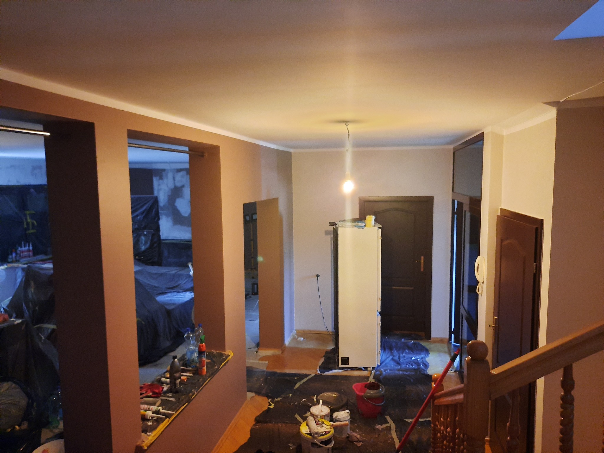 Wnętrze mieszkania w trakcie odświeżania: ściany pomalowane na dwa kolory, zabezpieczona podłoga folią, widoczne narzędzia malarskie i drabina. W tle pomieszczenie zabezpieczone folią malarską.