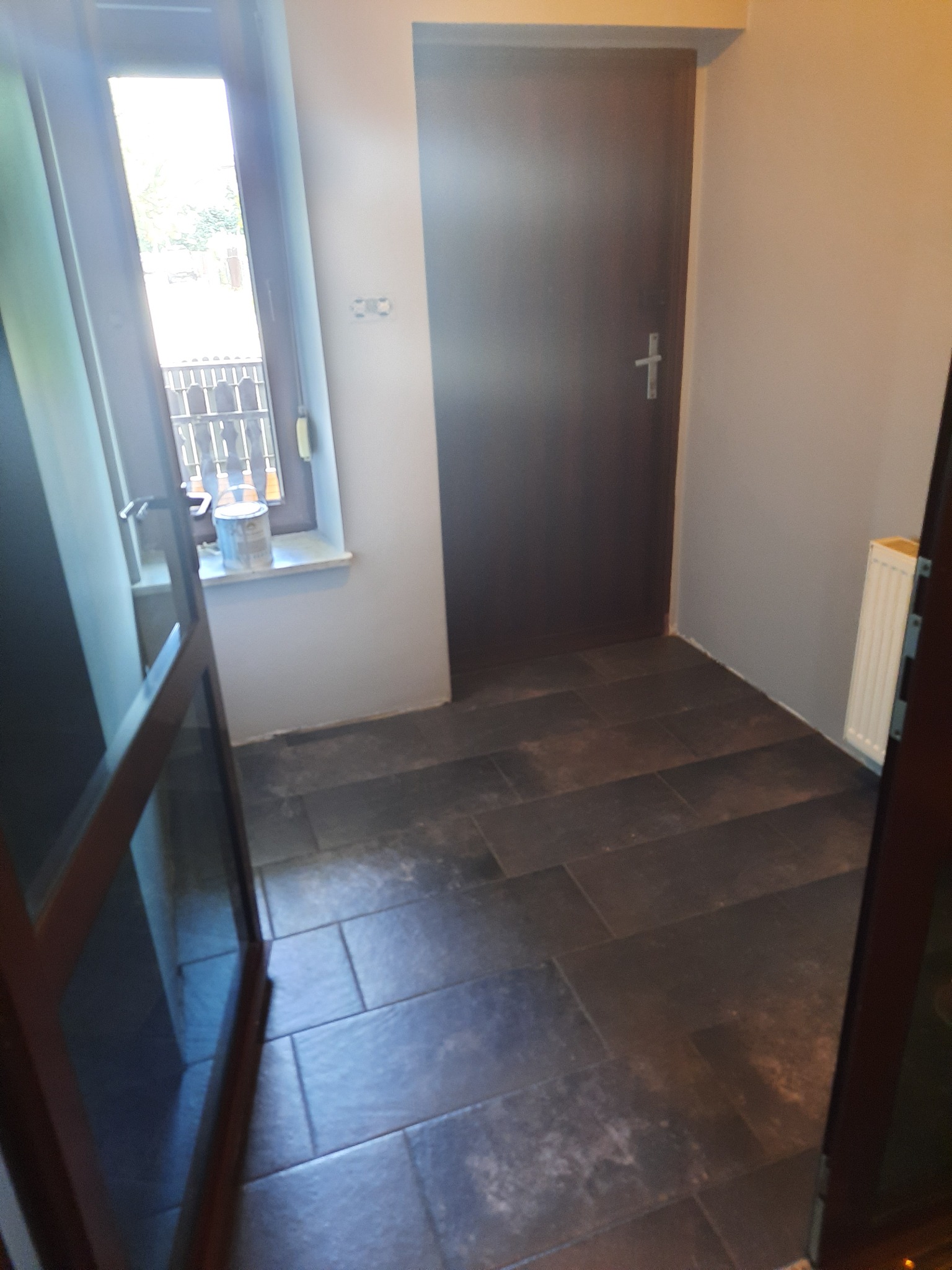 Wnętrze po remoncie: ciemnoszare płytki podłogowe ułożone w prostokąty, widoczna ściana z grzejnikiem, drzwi wewnętrzne w kolorze ciemnego brązu oraz okno z widokiem na zewnątrz, na parapecie...