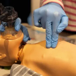 Ręce w niebieskich rękawiczkach wykonują resuscytację krążeniowo-oddechową na manekinie z użyciem worka ambu.