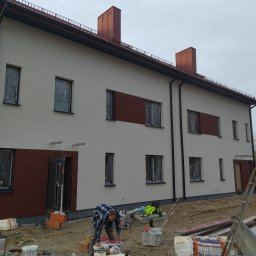 Kot-Bud - Pracownik w niebieskiej koszuli w kratę przygotowuje farbę na tle dwukondygnacyjnego budynku z jasną elewacją i brązowymi panelami, okna zabezpieczone folią, widoczne wiadra z farbą i narzędzia.