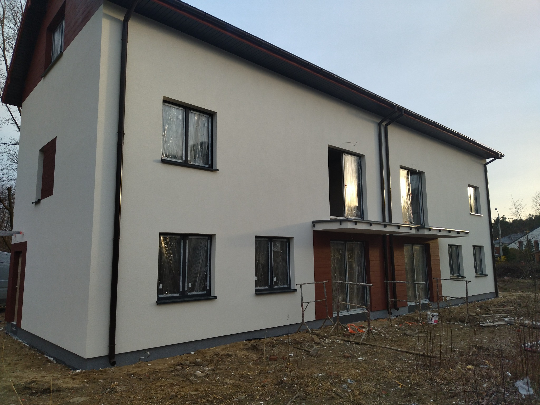 Dwupiętrowy budynek z nową, białą elewacją, czarnymi rynnami i oknami zabezpieczonymi folią, otoczony ziemią i rusztowaniami, w trakcie prac wykończeniowych.