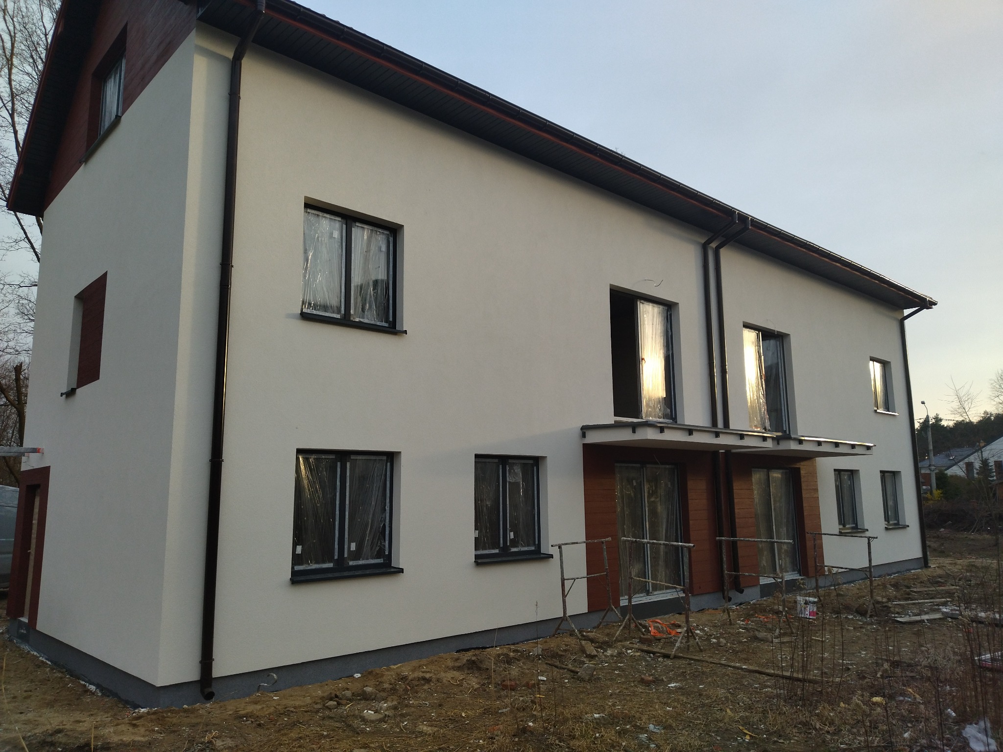 Dwupiętrowy budynek z białą elewacją, czarnymi oknami zabezpieczonymi folią i brązowym wykończeniem dachu, w trakcie prac wykończeniowych, z widocznym przygotowanym terenem wokół budynku.