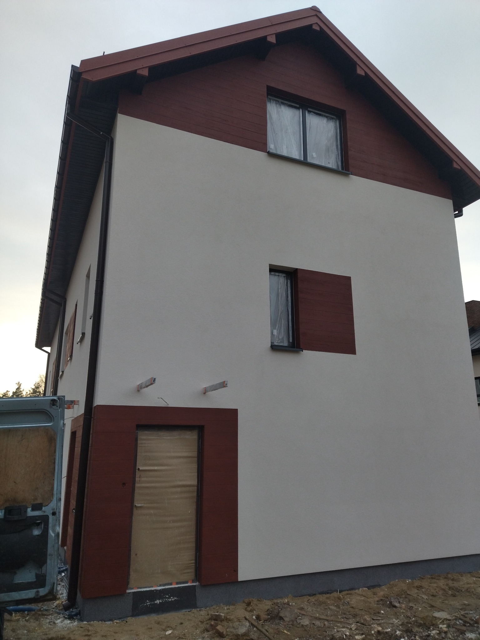 Dwukondygnacyjny dom z jasną elewacją i drewnianymi elementami wykończeniowymi w kolorze brązowym, okna zabezpieczone folią, widoczne ślady prac budowlanych.