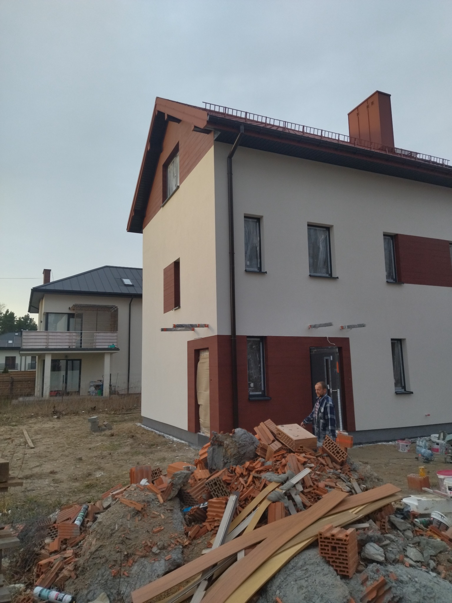 Dwukondygnacyjny dom z częściowo pomalowaną elewacją w kolorach bieli i brązu, okna zabezpieczone folią, na pierwszym planie sterta gruzu i materiałów budowlanych, mężczyzna w kraciastej koszuli...