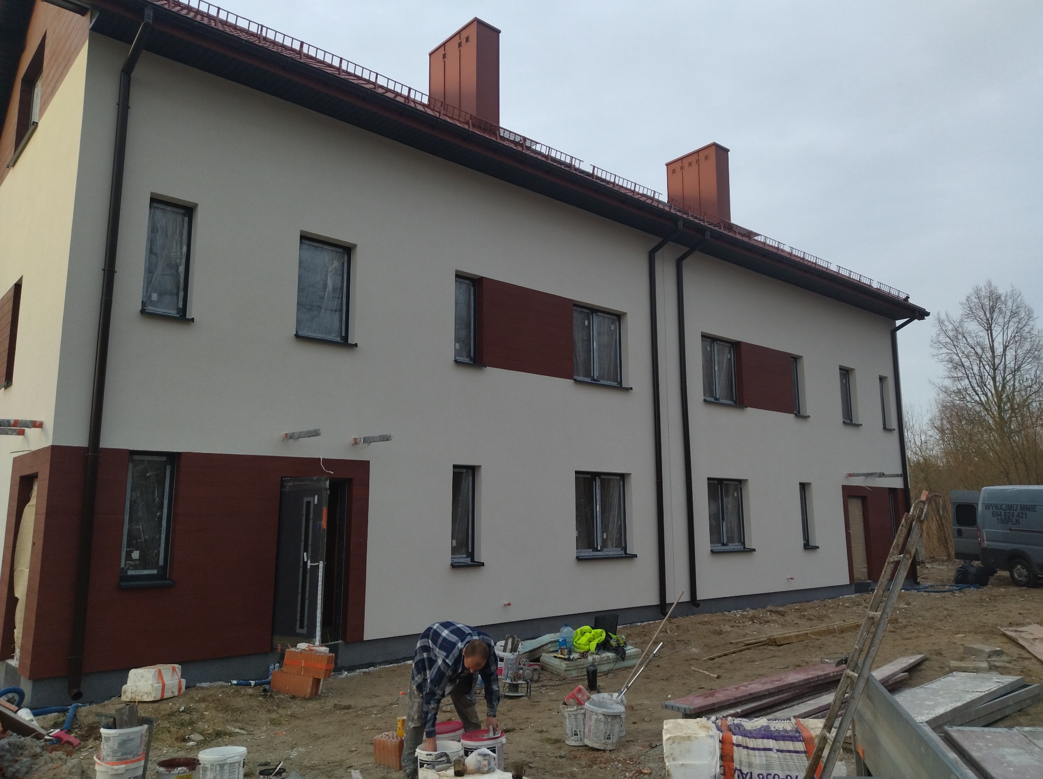 Pracownik w niebieskiej koszuli w kratę przygotowuje farbę na tle dwukondygnacyjnego budynku z jasną elewacją i brązowymi panelami, okna zabezpieczone folią, widoczne wiadra z farbą i narzędzia.