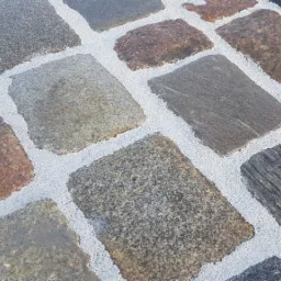 Ułożona kostka brukowa z różnokolorowego kamienia, wypełniona jasnym, drobnym kruszywem w spoinach.