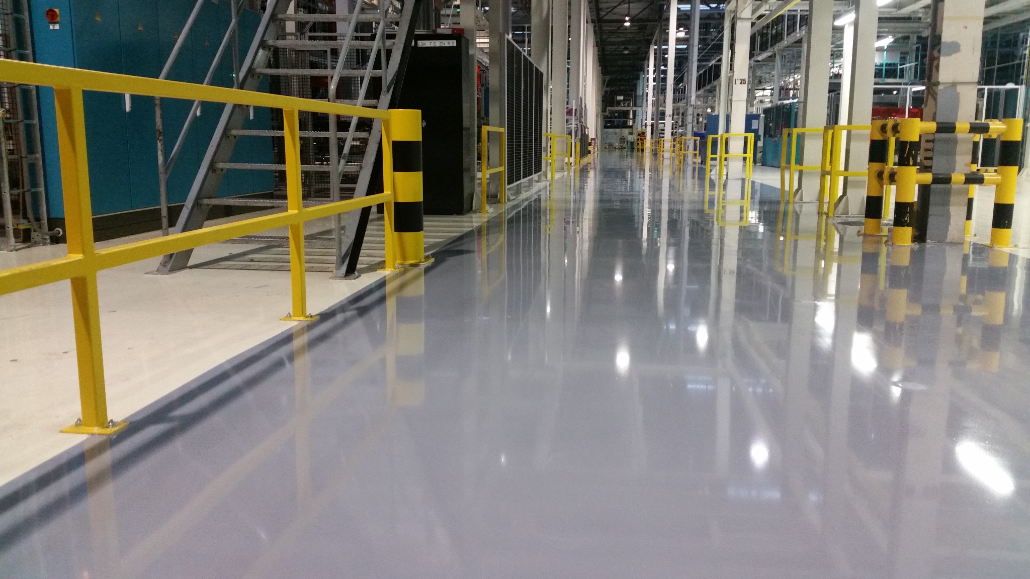 Gładka, szara posadzka w hali przemysłowej z żółtymi barierkami ochronnymi i czarno-żółtymi słupkami, odbijająca światło sufitu.