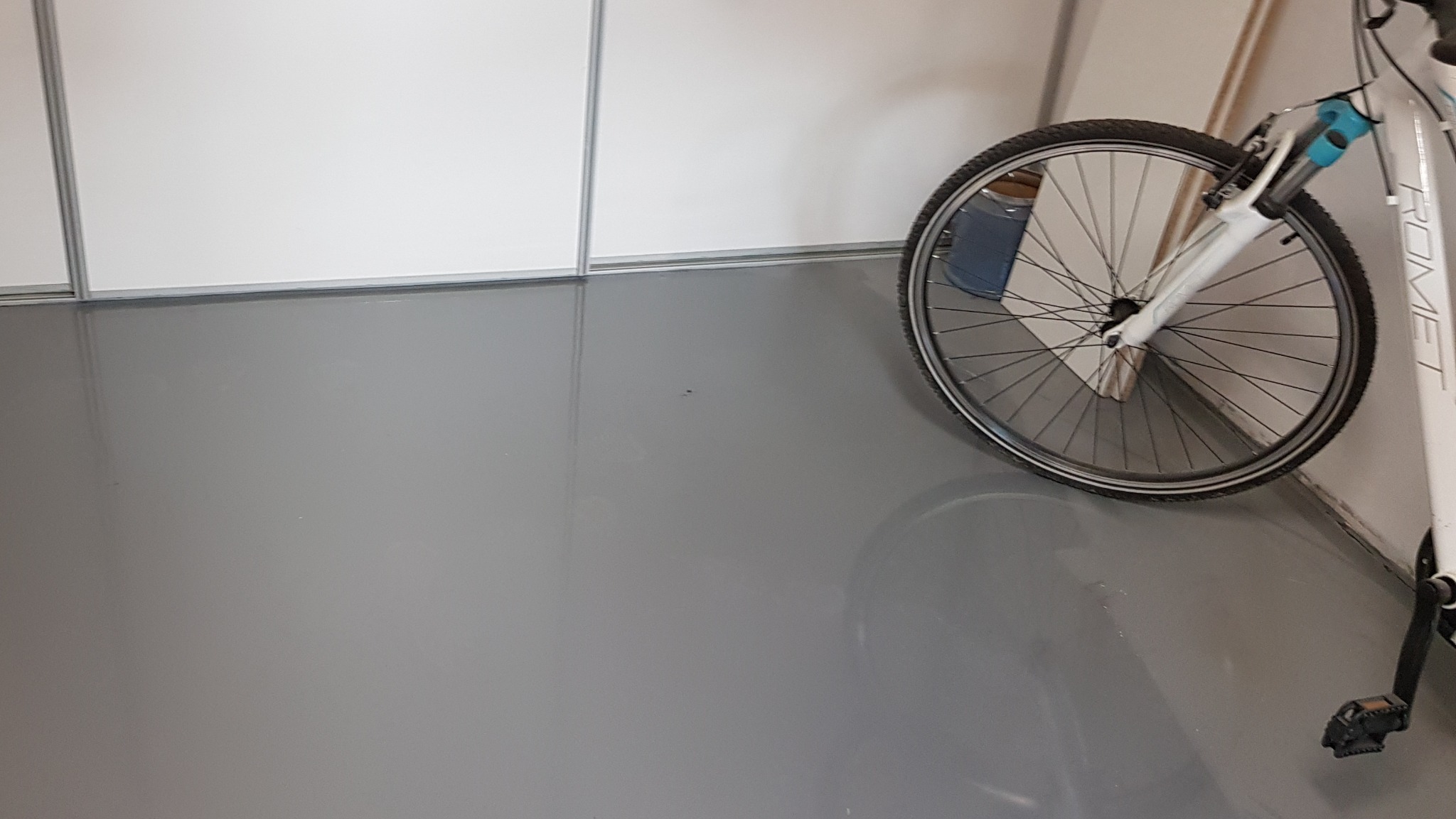 Gładka, szara powierzchnia podłogi z połyskiem, odbijająca częściowo rower z białym widelcem i czarną oponą, stojący przy białej ścianie.