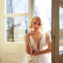 Portret kobiety z platynowymi włosami w białej sukience na ramiączkach, pozującej przy otwartym oknie z widokiem na drzewa.