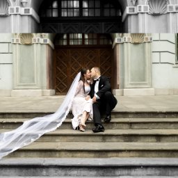 Karolina Pierścieniak Fotografia - Para młoda w czerni i bieli: Panna młoda w koronkowej sukni i welonie, pan młody w smokingu, siedzą na schodach i całują się na tle zabytkowej architektury.