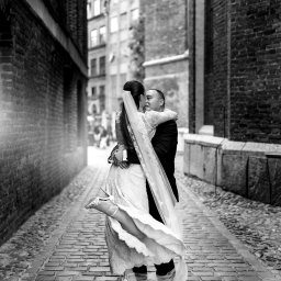 Karolina Pierścieniak Fotografia - Czarno-białe zdjęcie pary młodej w objęciach na brukowanej uliczce, panna młoda w koronkowej sukni i welonie, unosi nogę w białym bucie. Tło z ceglanych budynków.