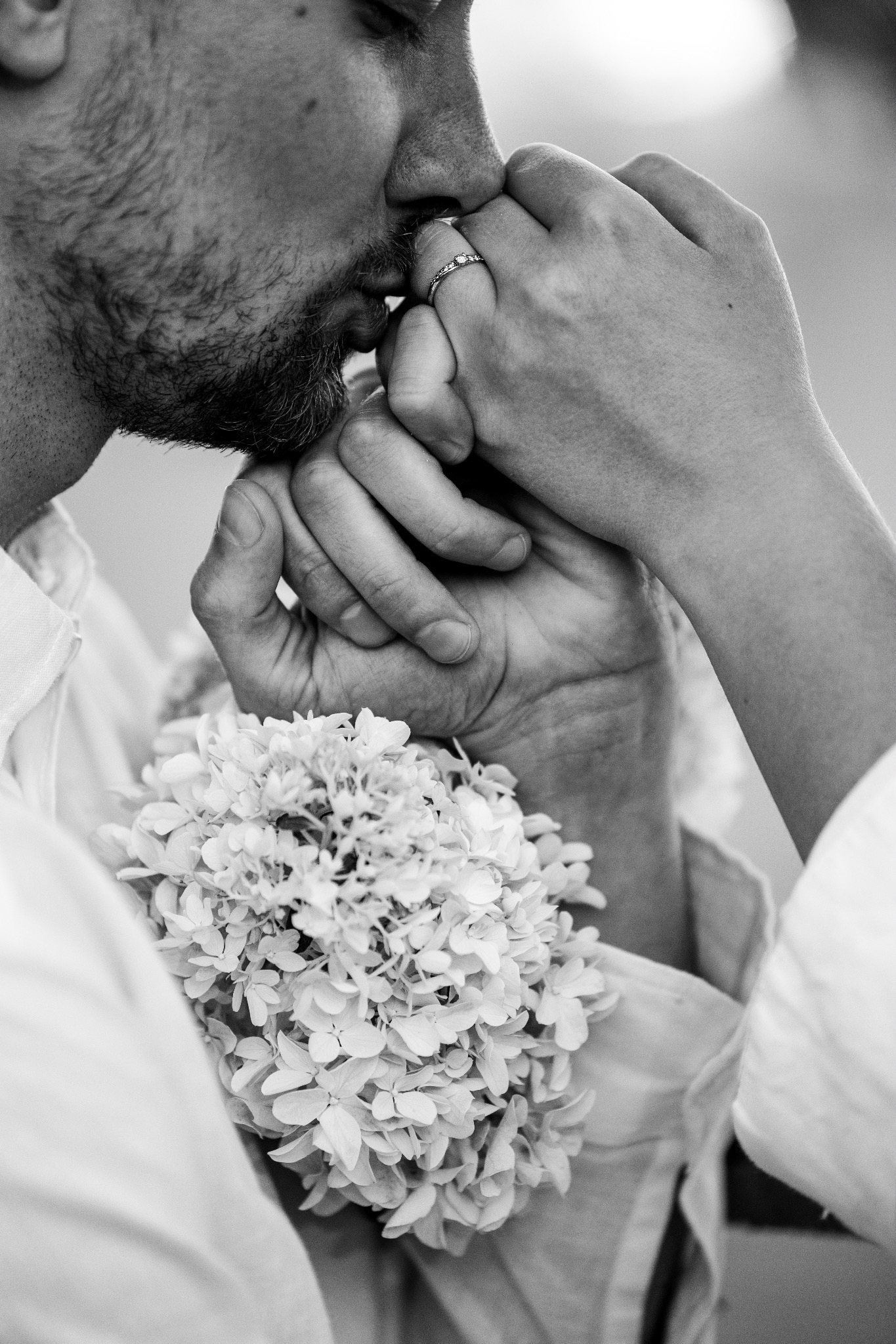 Czarno-białe zbliżenie: Mężczyzna całuje dłoń kobiety z pierścionkiem zaręczynowym, bukiet białych kwiatów przy koszuli.