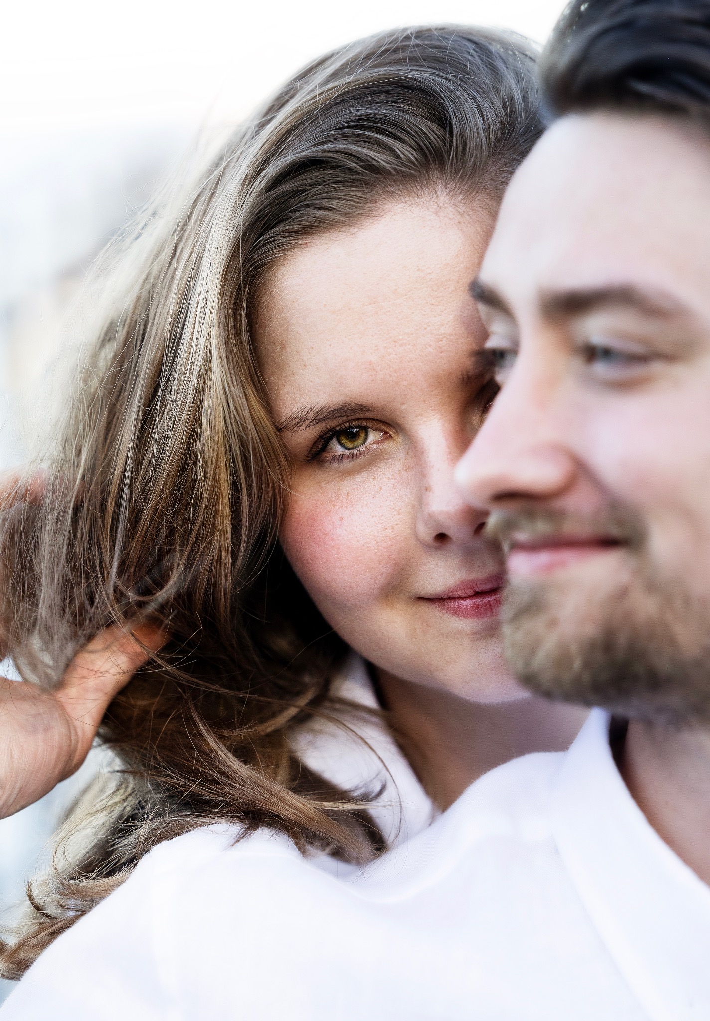 Portret pary: kobieta z zielonymi oczami spogląda w obiektyw, delikatnie dotykając włosów, obok niej w tle widoczny mężczyzna z brodą.