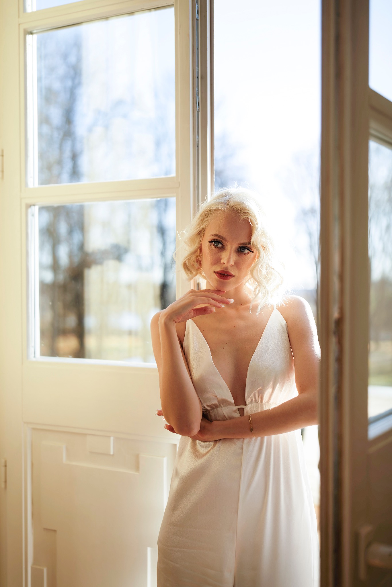 Portret kobiety z platynowymi włosami w białej sukience na ramiączkach, pozującej przy otwartym oknie z widokiem na drzewa.