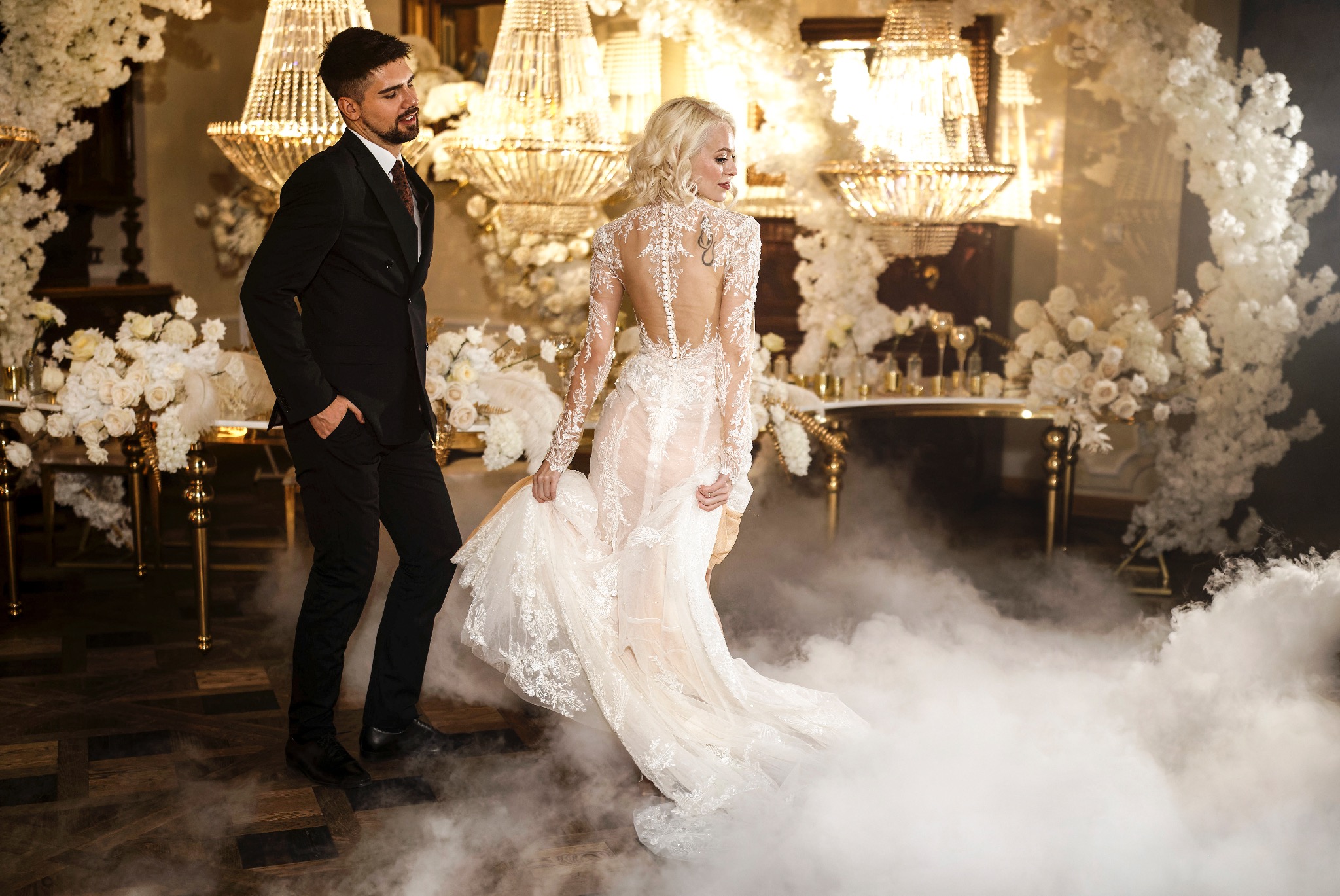 Elegancka para w otoczeniu dekoracji ślubnych i dymu: mężczyzna w czarnym garniturze, kobieta w koronkowej sukni z odkrytymi plecami, wzór tatuażu na plecach, kryształowe żyrandole i kompozycje...