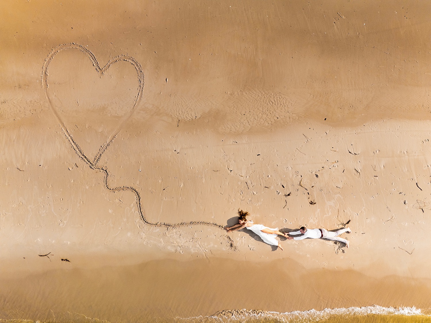 Para młoda leżąca na piasku obok wyrysowanego serca, widok z góry. Panna młoda w białej sukni, pan młody w białym garniturze. Delikatne fale obmywają brzeg.