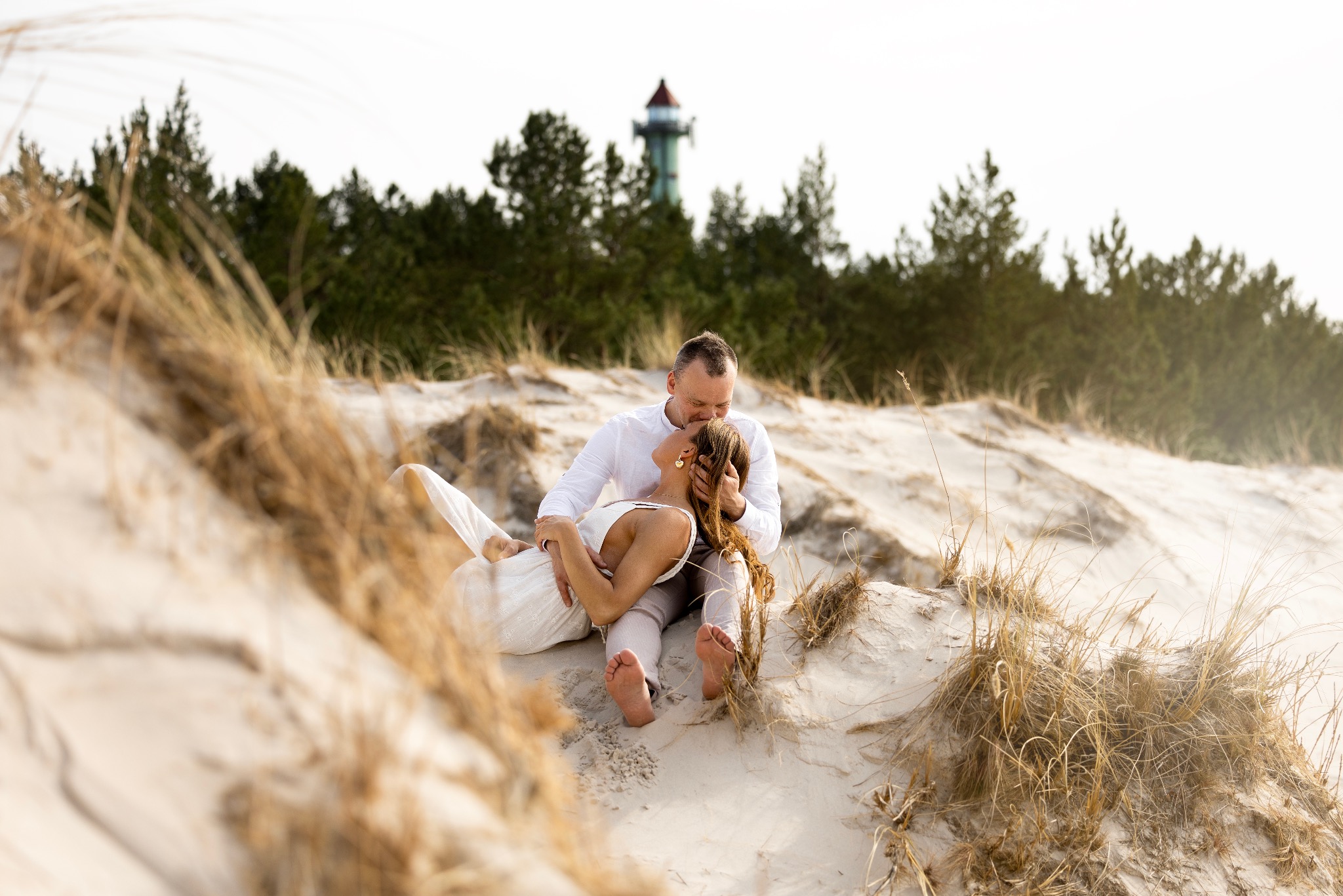Zakochana para na piaszczystej wydmie, mężczyzna całuje kobietę w czoło. W tle las i wieża latarni morskiej. Sesja narzeczeńska w naturalnej scenerii.