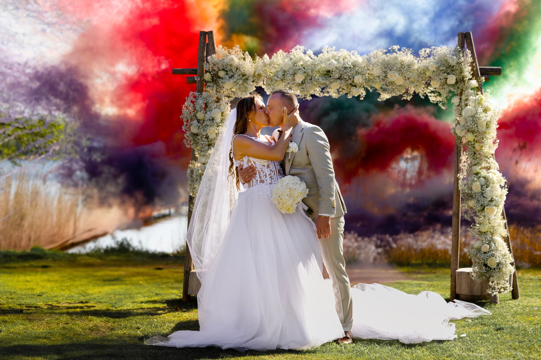 Pocałunek młodej pary pod drewnianym łukiem kwiatowym na tle kolorowego dymu. Panna młoda w białej sukni z bukietem, pan młody w beżowym garniturze. Romantyczna sceneria.