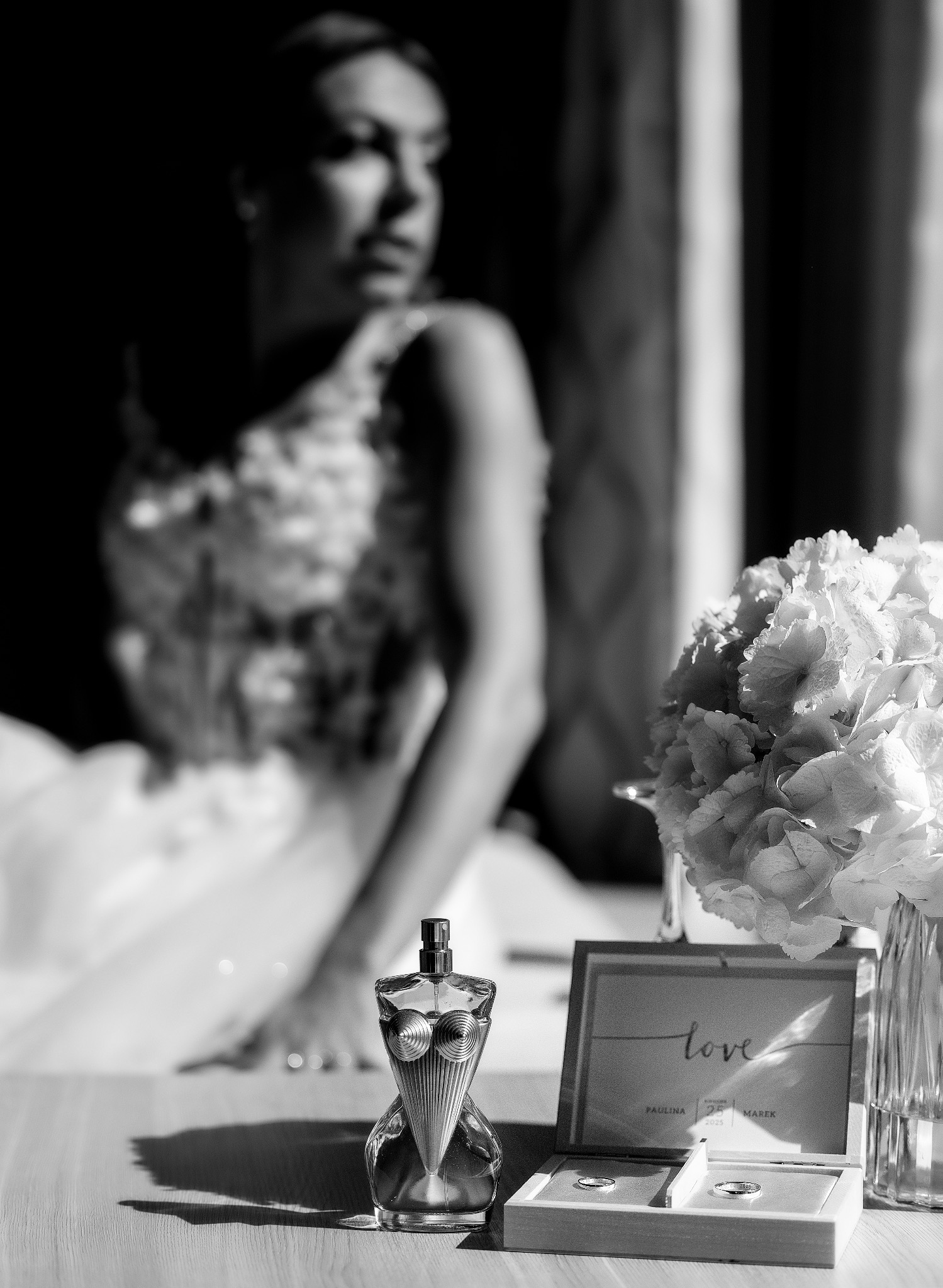 Czarno-białe zdjęcie ślubne: panna młoda w tle, flakon perfum, pudełko z obrączkami i bukiet kwiatów na pierwszym planie, subtelne światło.