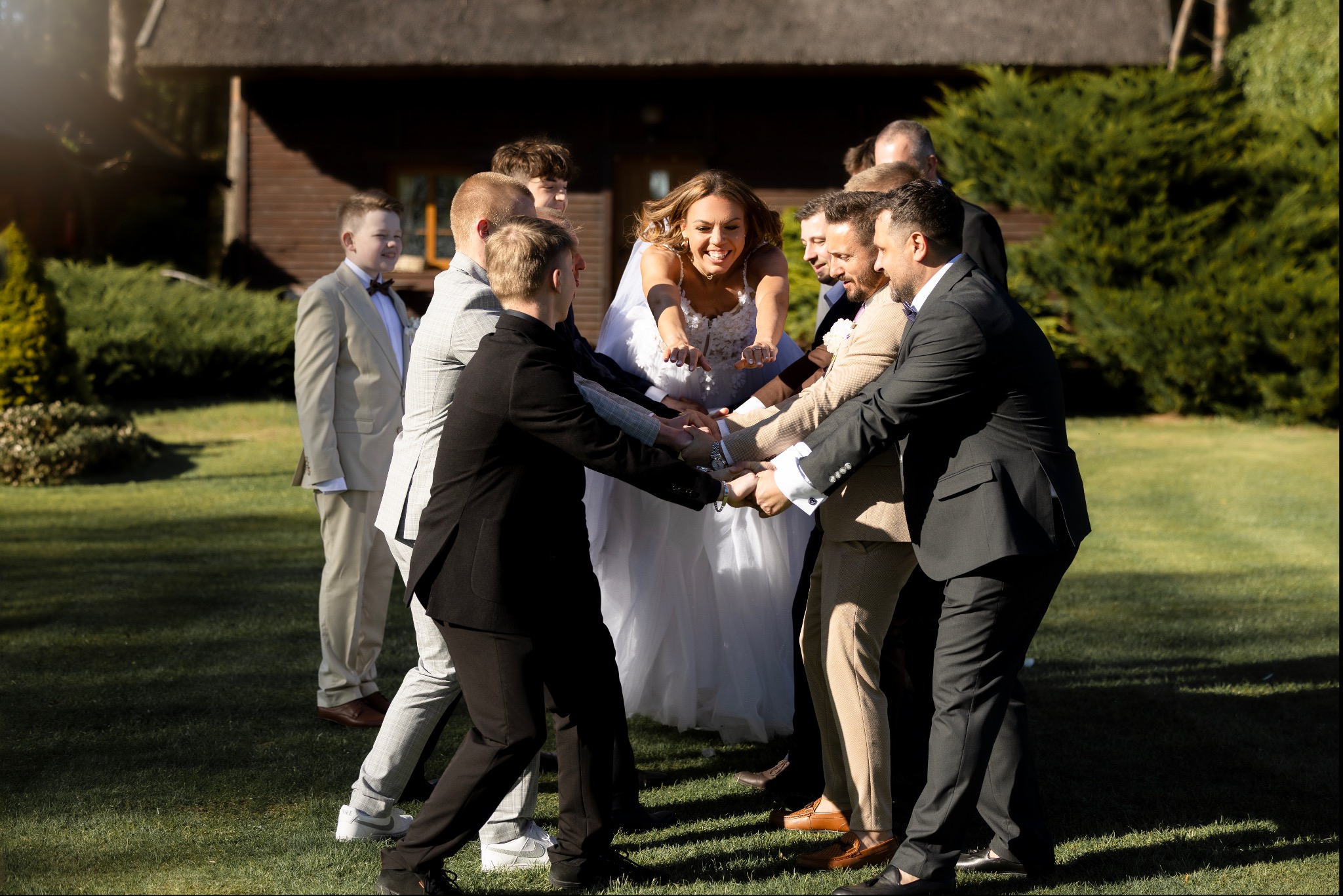 Radosna panna młoda w otoczeniu drużby, tworzących krąg z dłońmi na tle drewnianego budynku i zielonego trawnika w słoneczny dzień. Weselne ujęcie.