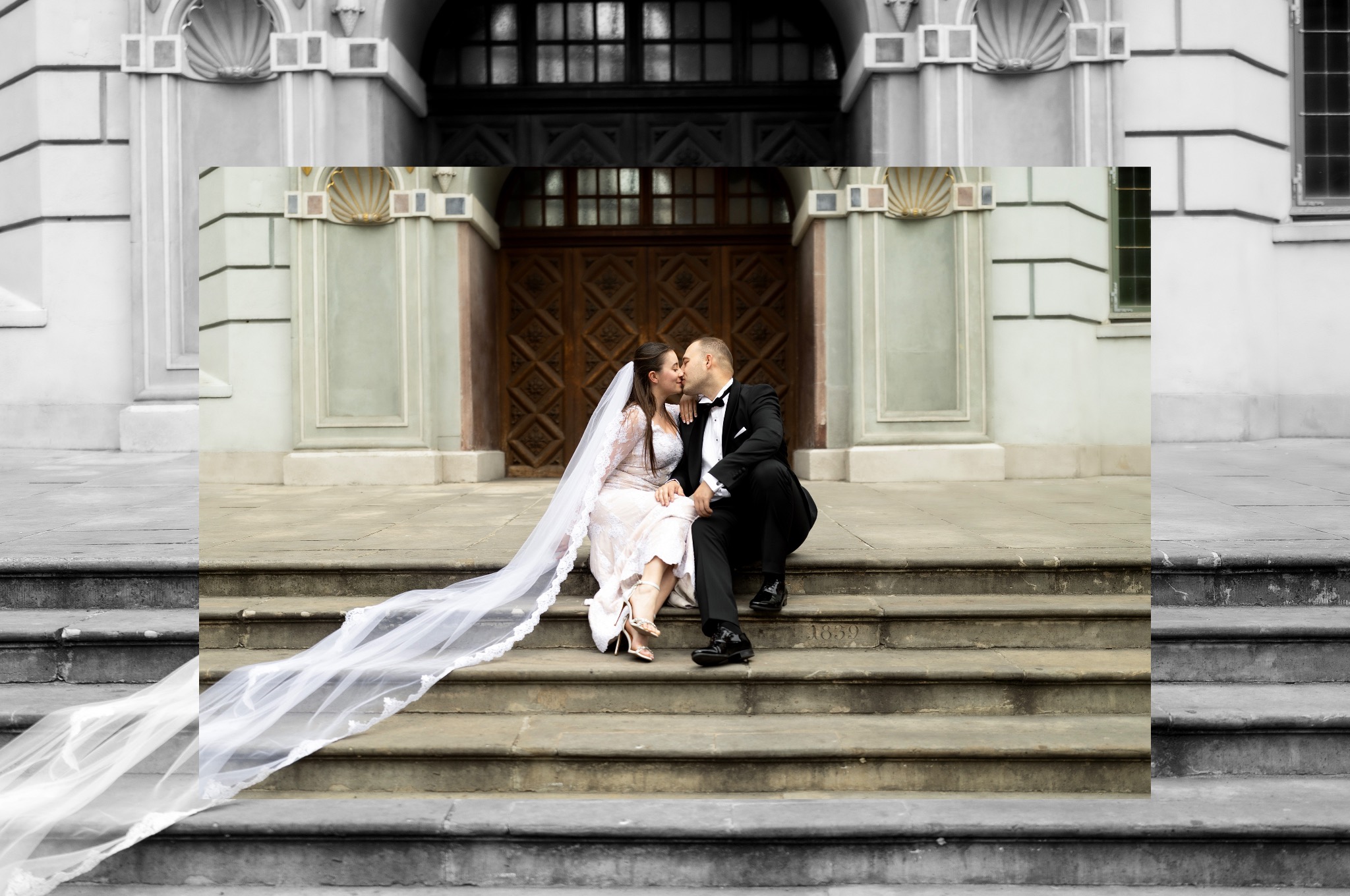 Para młoda w czerni i bieli: Panna młoda w koronkowej sukni i welonie, pan młody w smokingu, siedzą na schodach i całują się na tle zabytkowej architektury.