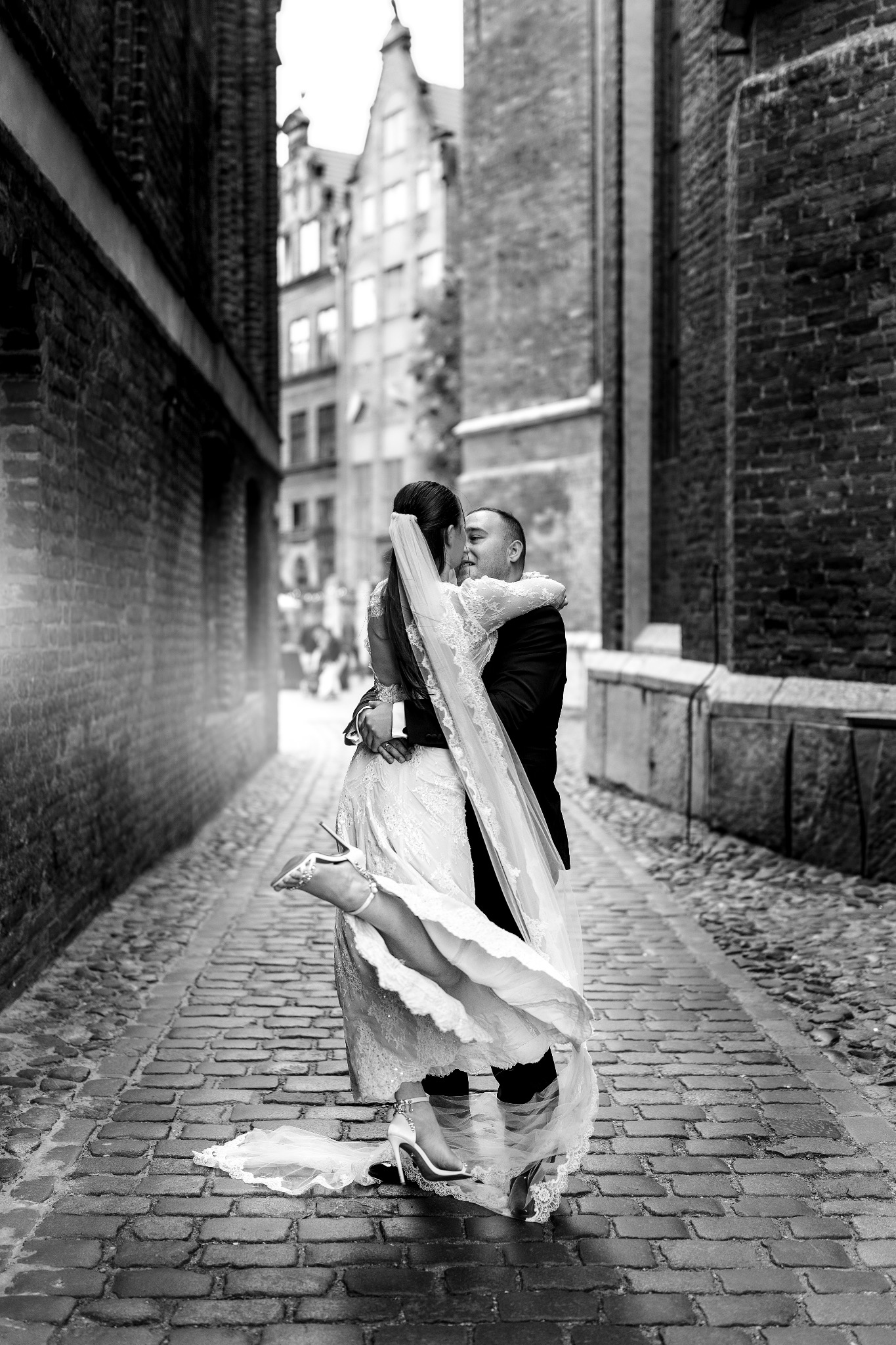 Czarno-białe zdjęcie pary młodej w objęciach na brukowanej uliczce, panna młoda w koronkowej sukni i welonie, unosi nogę w białym bucie. Tło z ceglanych budynków.