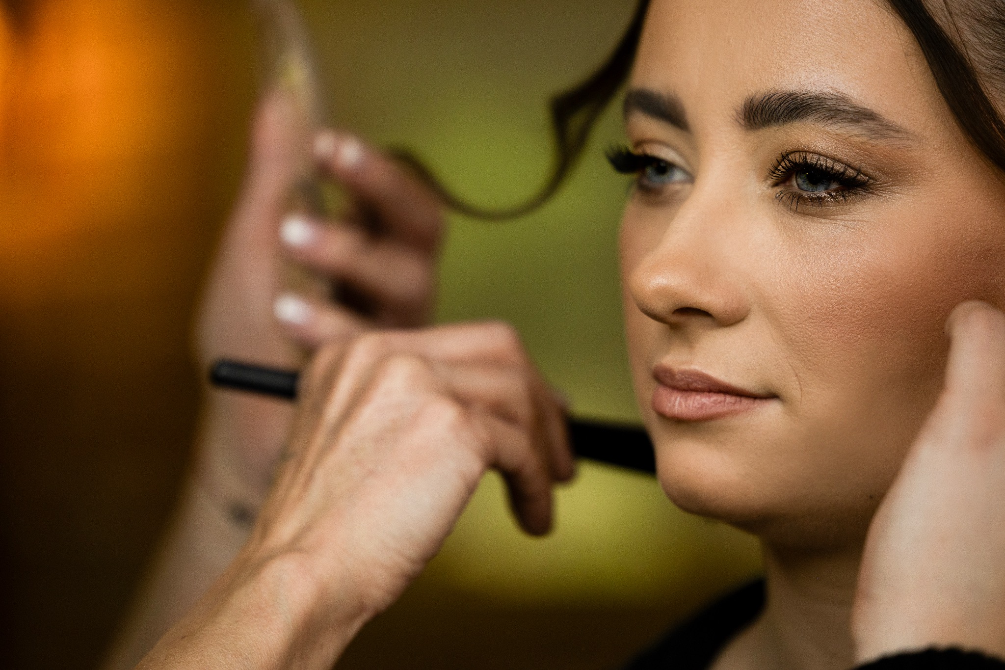 Portret kobiety w trakcie makijażu, z wyraźnie podkreślonym okiem i delikatnym uśmiechem, w tle dłoń wizażysty z pędzlem. Uwagę przykuwa intensywność spojrzenia.