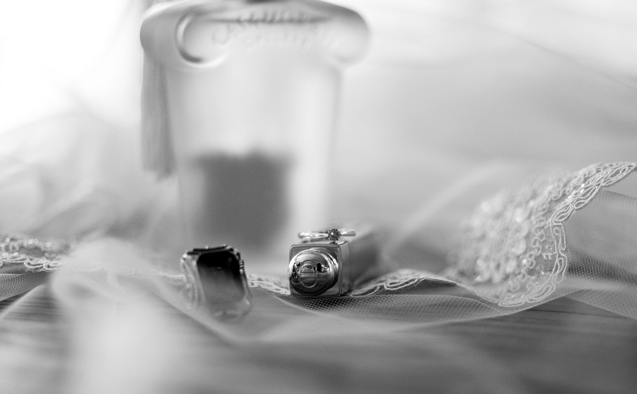 Czarno-białe zbliżenie pierścionków zaręczynowych, flakonu perfum i fragmentu welonu ślubnego z koronkowym wykończeniem, tworzące subtelną kompozycję.