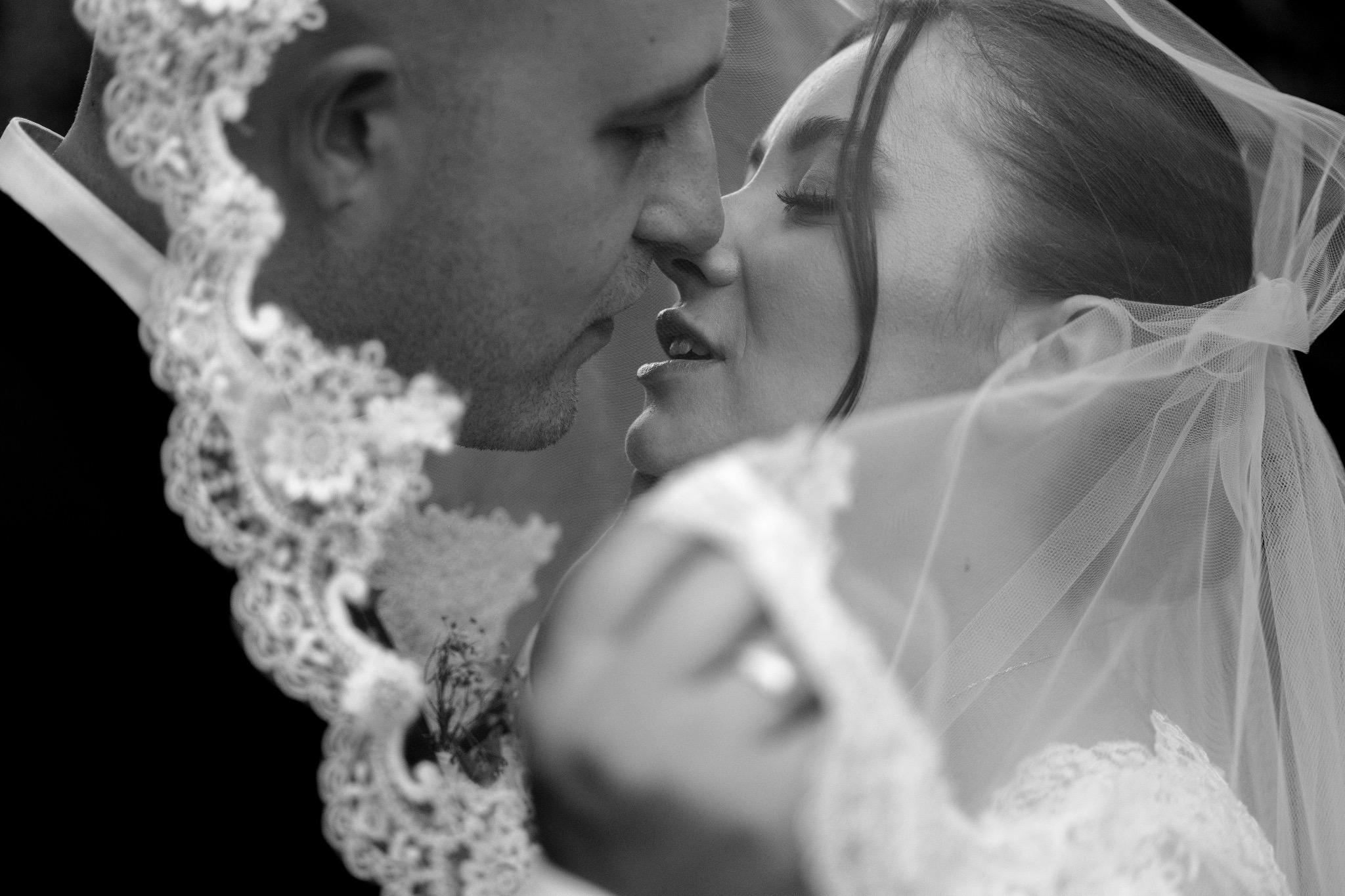 Czarno-białe zbliżenie pary młodej pod welonem, delikatny pocałunek, zamknięte oczy panny młodej, widoczne detale koronki welonu i sukni. Romantyczny moment, subtelne światło.