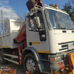 Biała ciężarówka Iveco z dźwigiem F65 i pomarańczowym osprzętem na przodzie, stojąca na piaszczystym podłożu, z kurami spacerującymi w pobliżu kół.