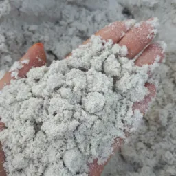 Dłoń trzymająca jasny, drobny materiał sypki, prawdopodobnie do wykorzystania w budownictwie lub ogrodnictwie, z pomalowanym na pomarańczowo paznokciem widocznym w lewym górnym rogu.