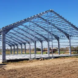 Stalowa konstrukcja szkieletowa budynku usługowego na betonowej płycie fundamentowej, widok z zewnątrz, w tle pola i zabudowania wiejskie, na pierwszym planie ziemia i fragment ciągnika.