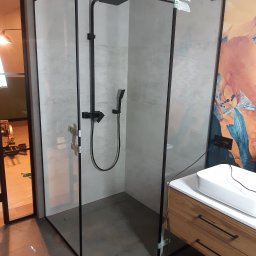 Kamil Olaf - Nowoczesna kabina prysznicowa z czarnymi ramami i deszczownicą, widoczne naklejki zabezpieczające na szkle, obok umywalka z drewnianą szafką.