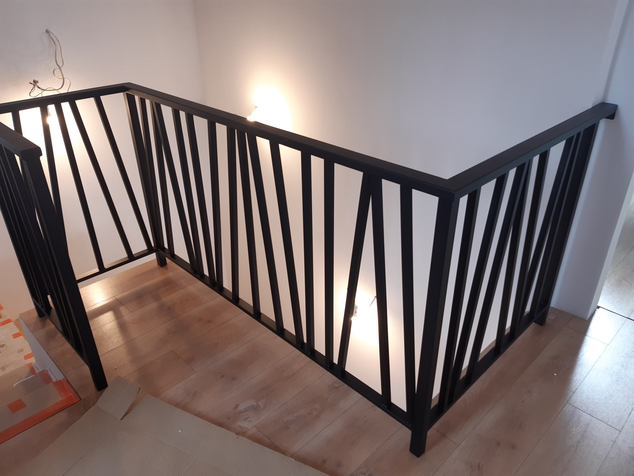 Nowoczesna, czarna balustrada schodowa z pionowymi, skośnymi prętami, zamontowana wewnątrz budynku z drewnianymi schodami i podłogą, widoczne niedokończone prace wykończeniowe.