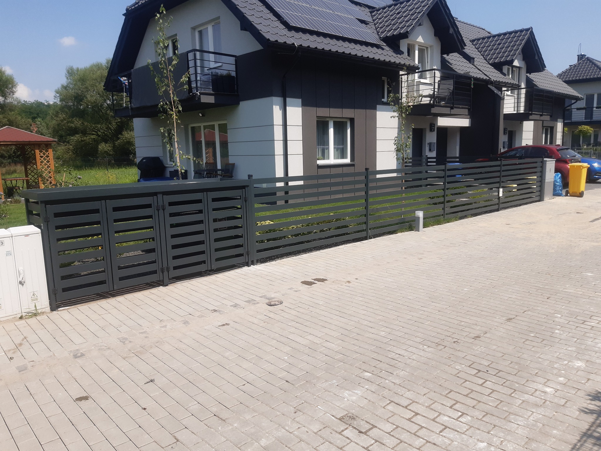 Nowoczesne ogrodzenie panelowe w kolorze grafitowym, z poziomymi przęsłami, oddzielające posesję z domami jednorodzinnymi o nowoczesnej architekturze, widoczne panele słoneczne na dachu.