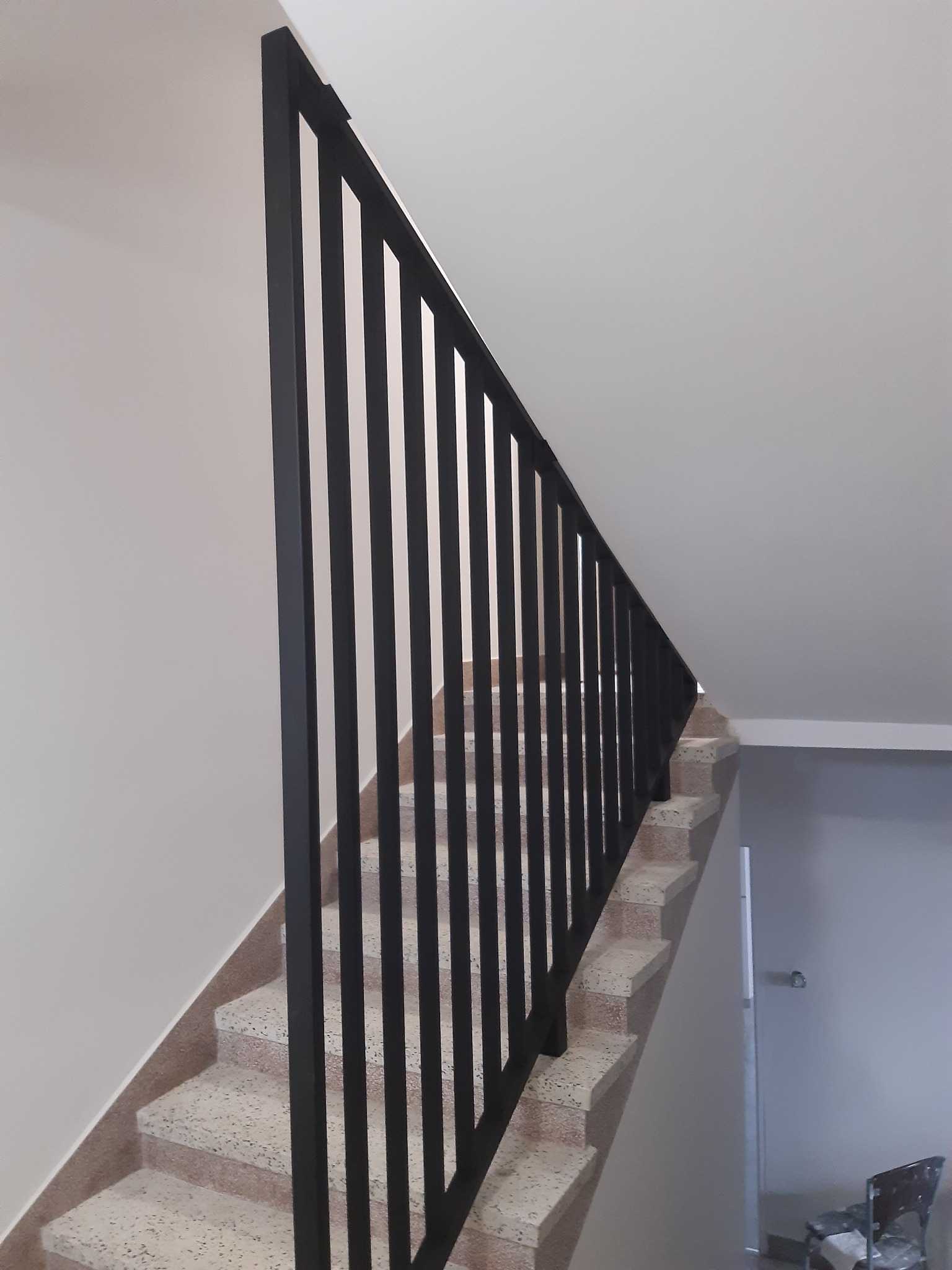 Minimalistyczna czarna balustrada z pionowych prętów zabezpieczająca schody z lastryko w jasnym wnętrzu.