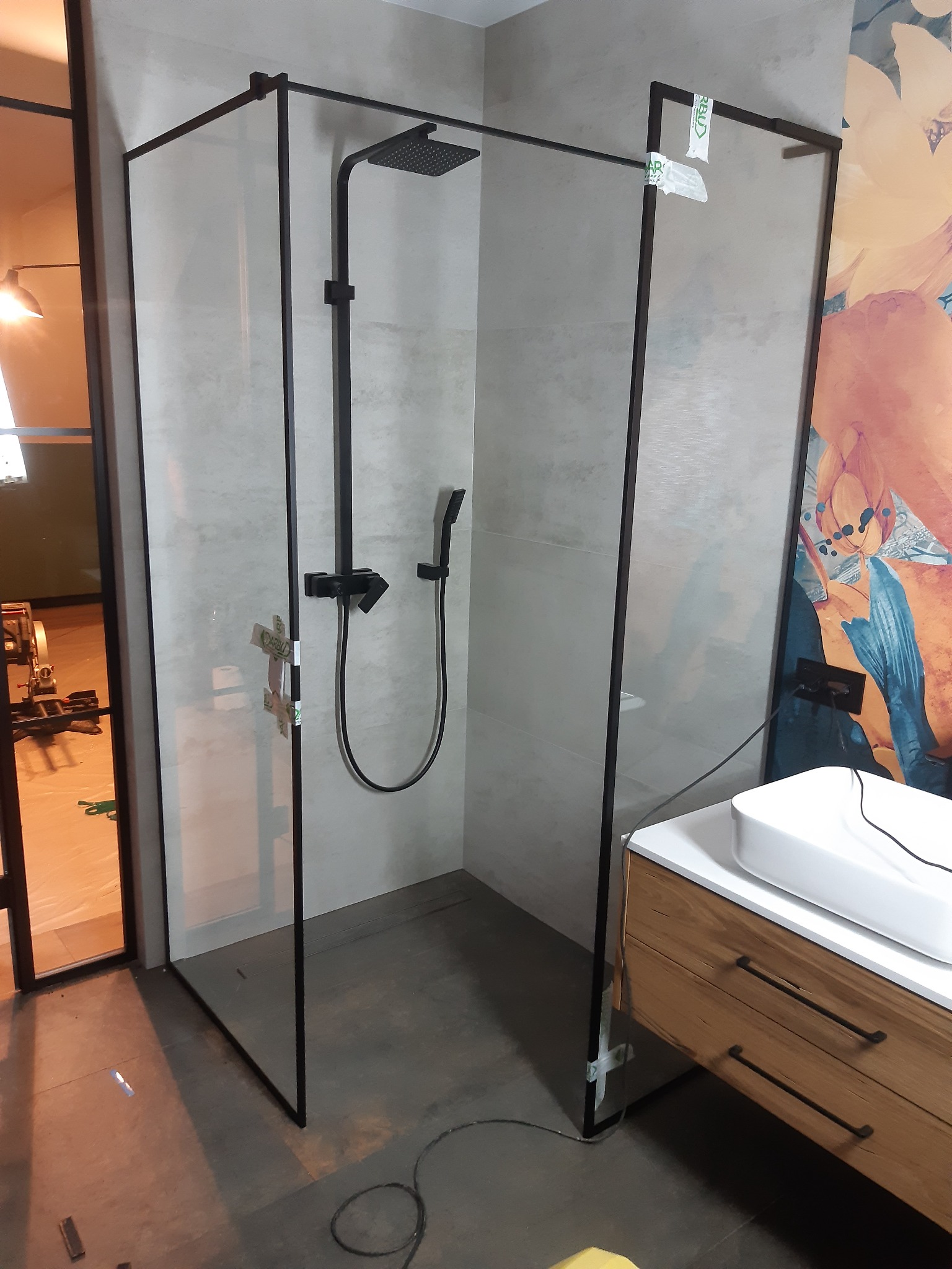 Nowoczesna kabina prysznicowa z czarnymi ramami i deszczownicą, widoczne naklejki zabezpieczające na szkle, obok umywalka z drewnianą szafką.