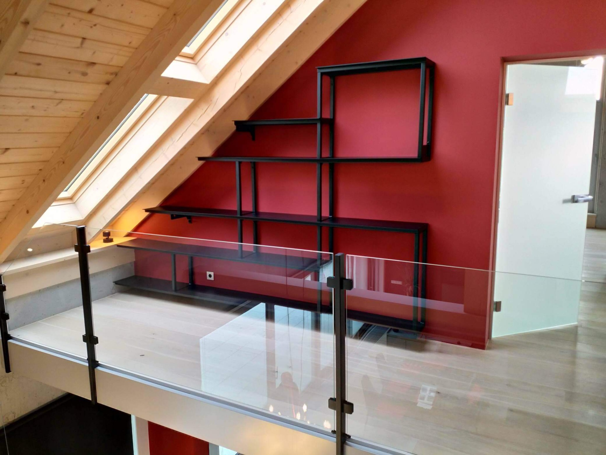 Wnętrze poddasza z przeszkloną balustradą, drewnianym sufitem z oknami dachowymi oraz metalowym regałem na czerwonej ścianie.