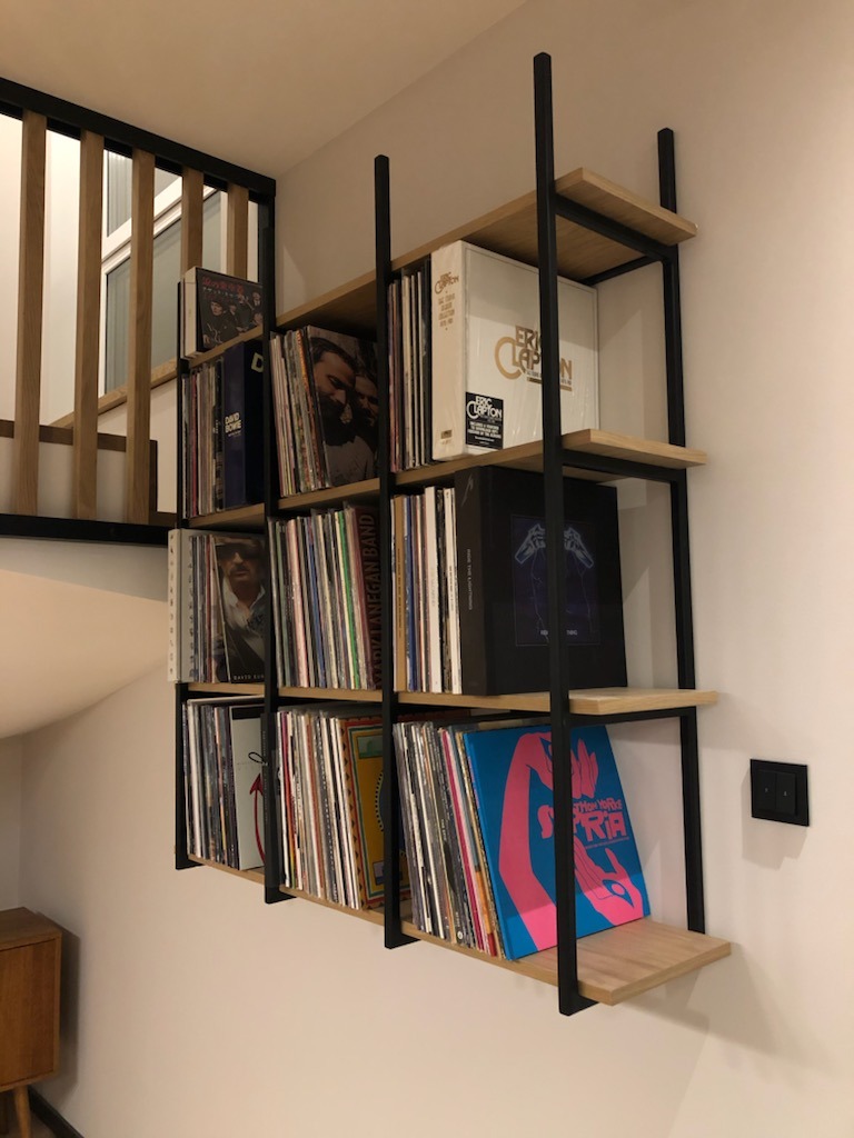 Regał wiszący z metalową ramą i jasnymi drewnianymi półkami, wypełniony płytami winylowymi, umieszczony na białej ścianie obok schodów z drewnianą balustradą.