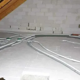 Instalacja przewodów wentylacyjnych w izolowanej poddaszowej przestrzeni, widoczne rury rozprowadzone po podłodze, elementy montażowe i fragment drewnianej konstrukcji dachu.