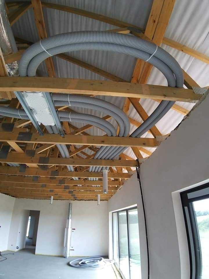 Montaż elastycznych przewodów wentylacyjnych pod drewnianą konstrukcją dachu w surowym wnętrzu budynku, widoczne rury spiro i skrzynka rozdzielcza.