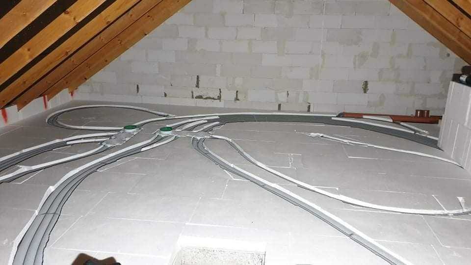 Instalacja przewodów wentylacyjnych w izolowanej poddaszowej przestrzeni, widoczne rury rozprowadzone po podłodze, elementy montażowe i fragment drewnianej konstrukcji dachu.