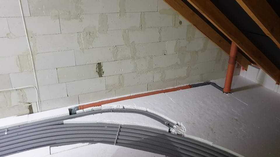 Szare, karbowane rury wentylacyjne ułożone na białej piance izolacyjnej poddasza, widoczne fragmenty ścian z białego bloczka, pomarańczowe rury pionowe i poziome, oraz drewniana konstrukcja dachu.