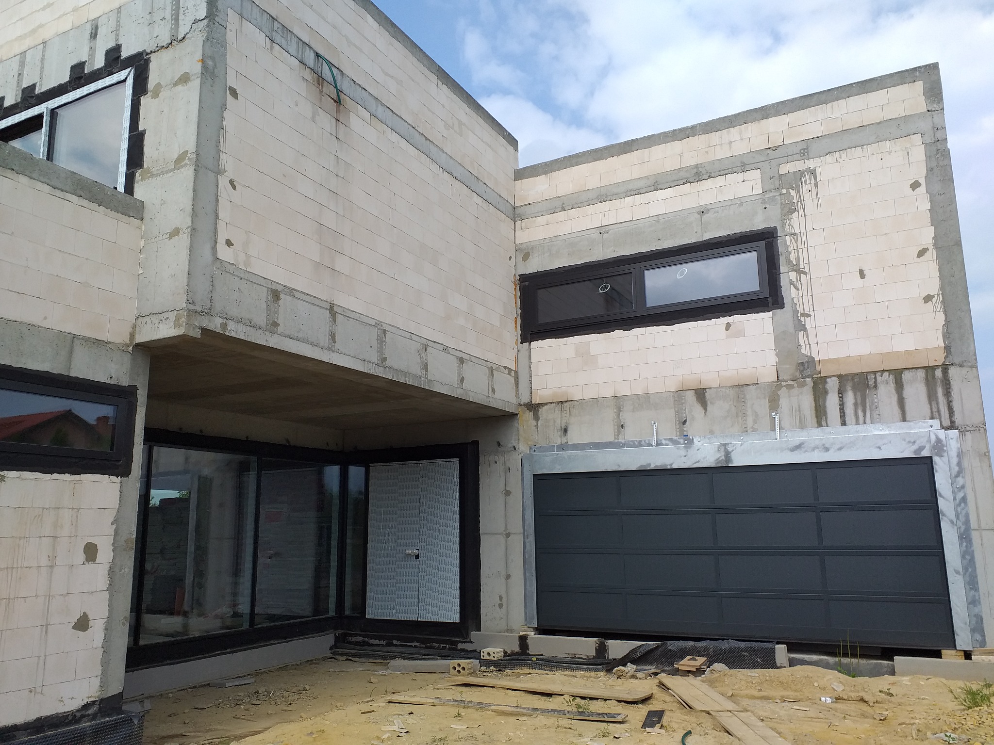 Surowy stan nowoczesnego domu z betonu komórkowego, widoczne okna z czarnymi ramami, brama garażowa w trakcie montażu, teren budowy z piaskiem i elementami konstrukcyjnymi.
