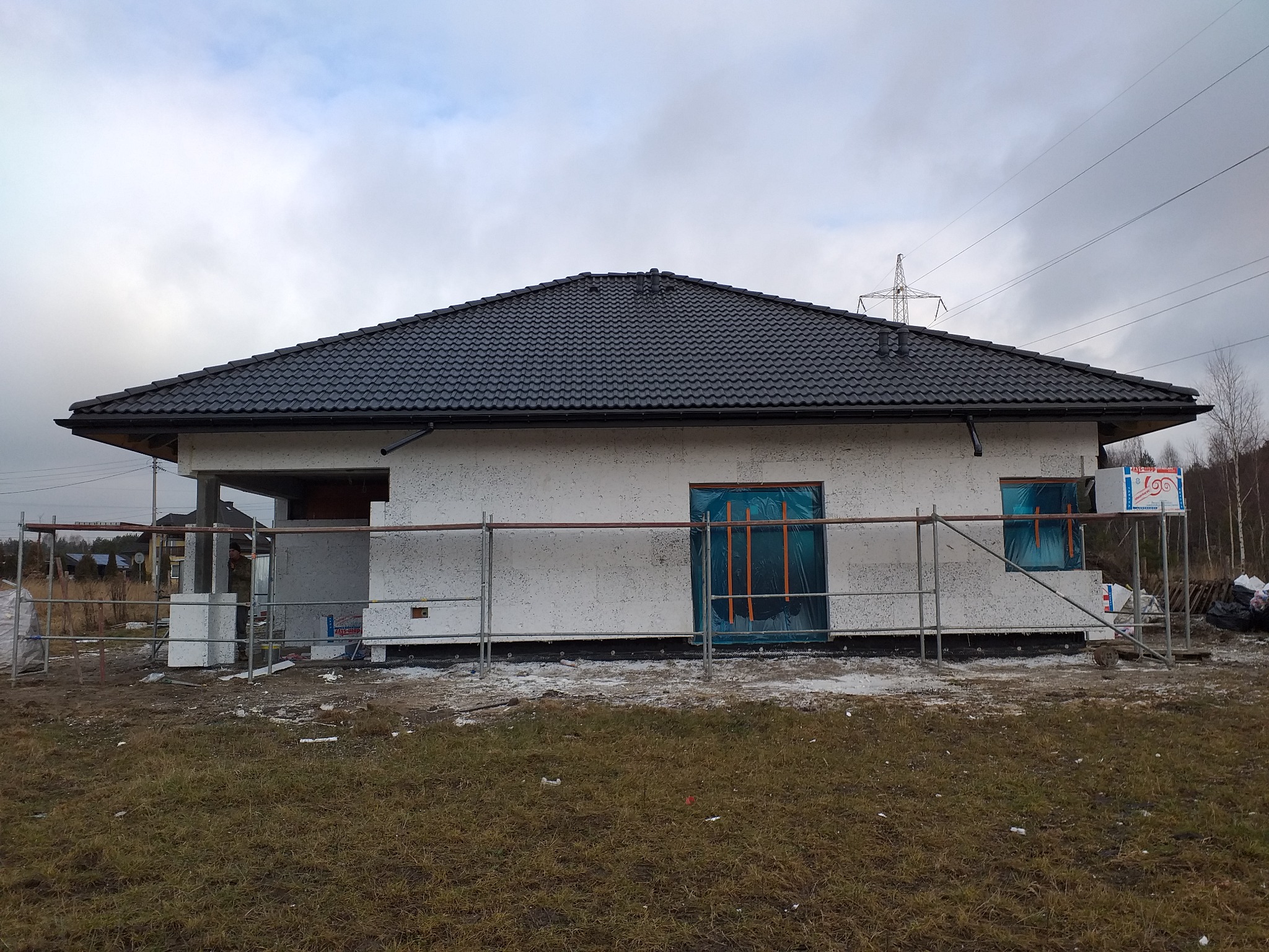 Dom murowany w trakcie budowy, z czarnym dachem z dachówki, zabezpieczone okna folią, widoczne rusztowania i teren budowy w pochmurny dzień.