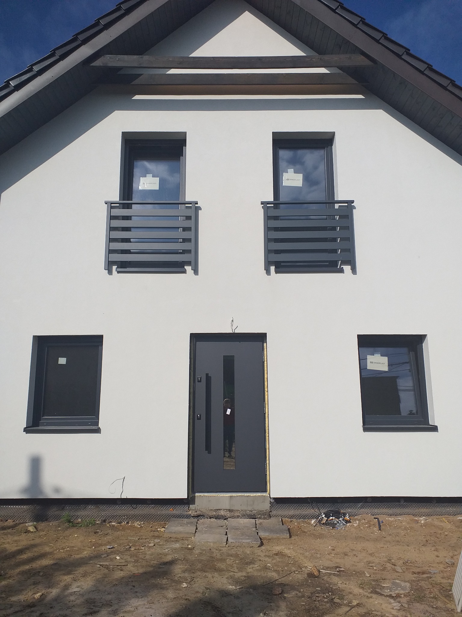 Nowoczesny dom z białymi ścianami, szarą stolarką okienną i drzwiami, oraz balkonami z poziomymi balustradami. Widoczne wystające kable przy fundamencie.