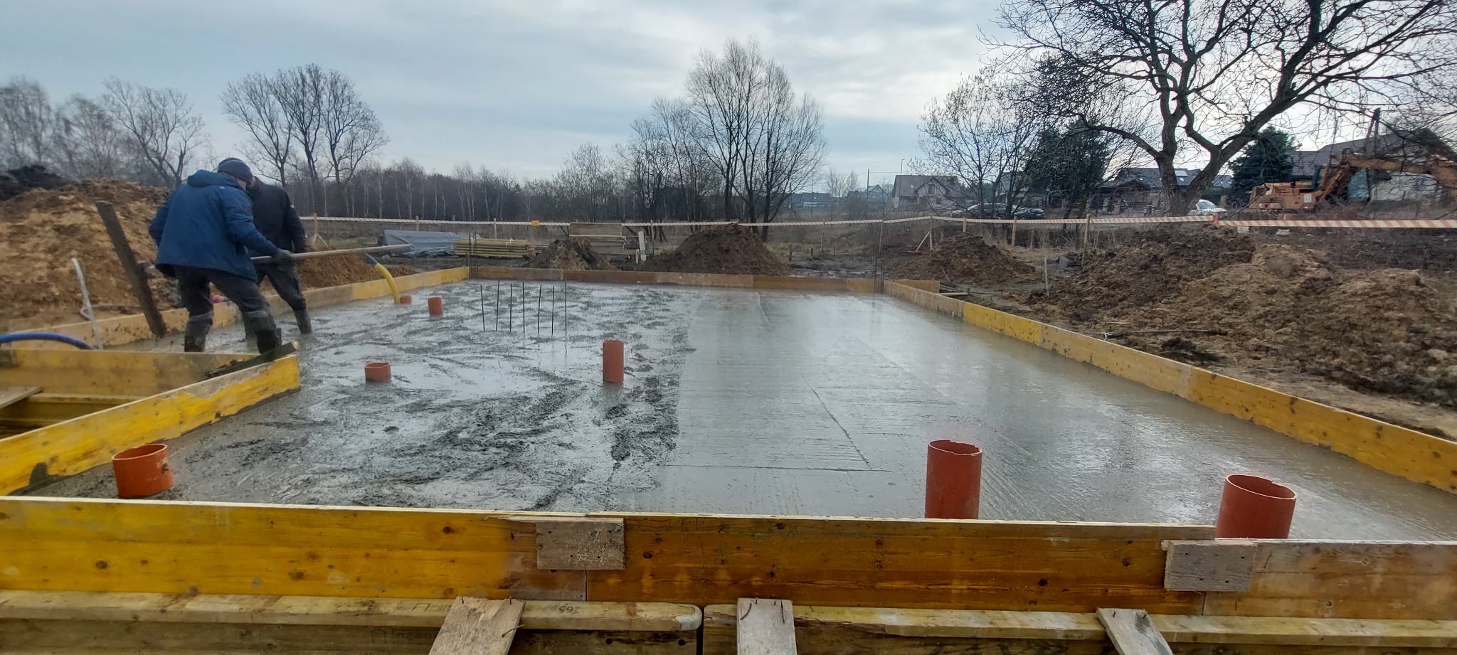 Wylewka fundamentowa z szalunkiem drewnianym i wystającymi rurami kanalizacyjnymi, w tle drzewa bez liści i częściowo widoczny budynek.