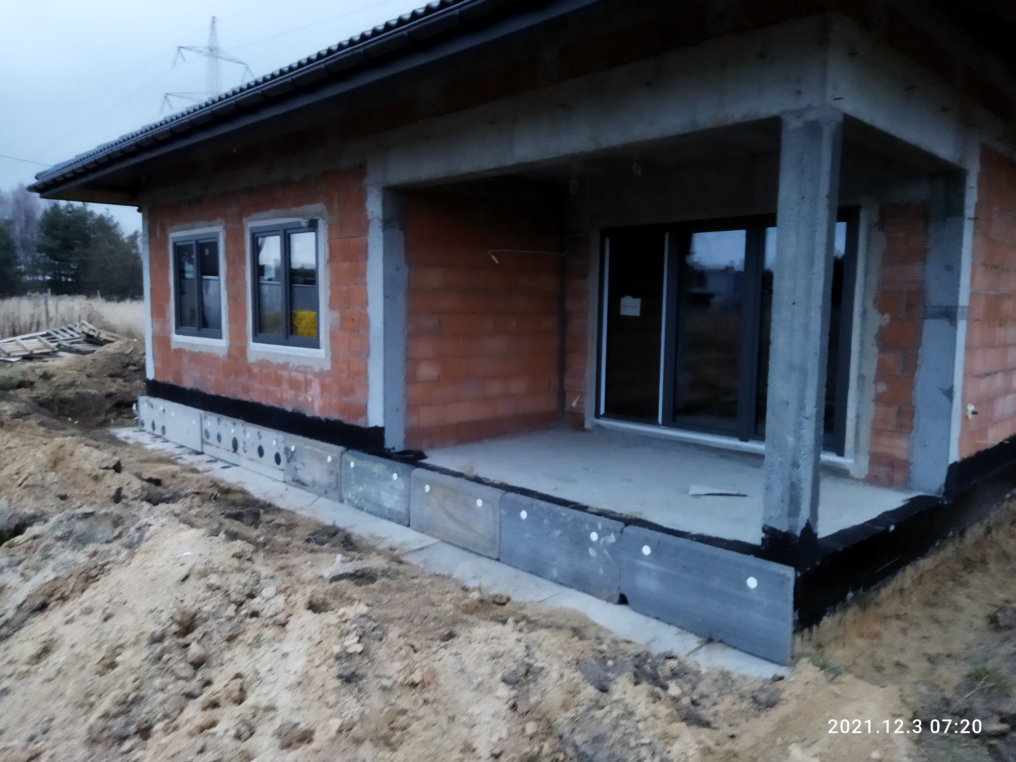 Surowy stan budowy parterowego domu z czerwonej cegły, widoczne szare okna, betonowe filary werandy i izolacja fundamentów, teren wokół w trakcie prac ziemnych, pochmurne niebo.